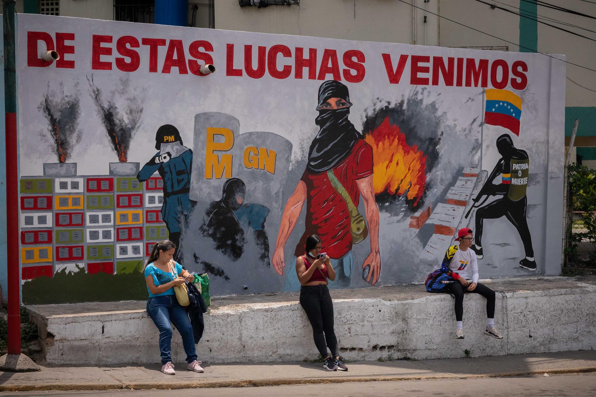 Varias personas esperan frente a un mural que retrata algunos hechos de manifestaciones y violencia relacionada a la política en Venezuela. Foto: EFE